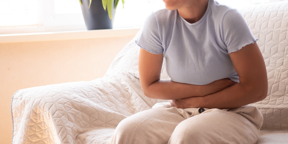 Vrouw in een beige broek en een lichtblauw t-shirt die met haar handen voor haar buik zit omdat ze last heeft van chronische buik klachten