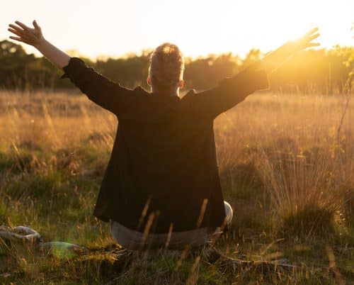 Jolanda Pouwels zit in een veld met uitgestrekte armen richting de zonsondergang, genietend van de rust en natuur.