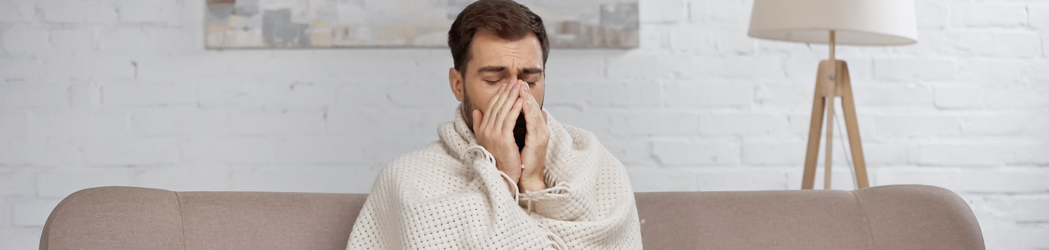 Afbeelding van een man die op de bank zit met een deken om zich heen die duidelijk ziek is en met zijn handen voor zijn mond zit. Achter hem staat een lamp.