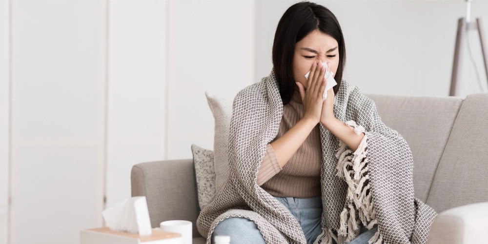 Foto van een aziatische vrouw met een deken om haar schouders. Ze zit op een lichtbeige bank en snuit het snot uit haar neus met een zakdoek omdat ze verkouden en niet fit is.