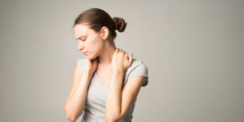 Een afbeelding van een vrouw met een beige tshirt aan die haar handen op de pijnlijke spieren van haar nek heeft liggen. Ze wil weten hoe ze fibromyalgie kan ontdekken