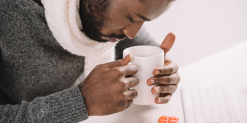 Afbeelding van een man die ziek is. Hij drinkt een kop boullion en heeft pillen voor zich ligen. Hij weet nog niet dat het lichaam niet tegen je werkt als je ziek bent